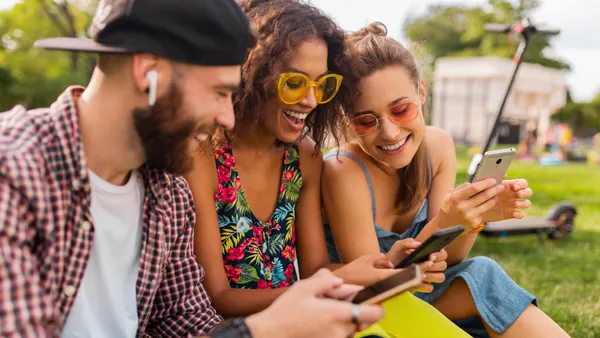 Three young adults sit on grass in a sunny park, smiling and looking at their smartphones