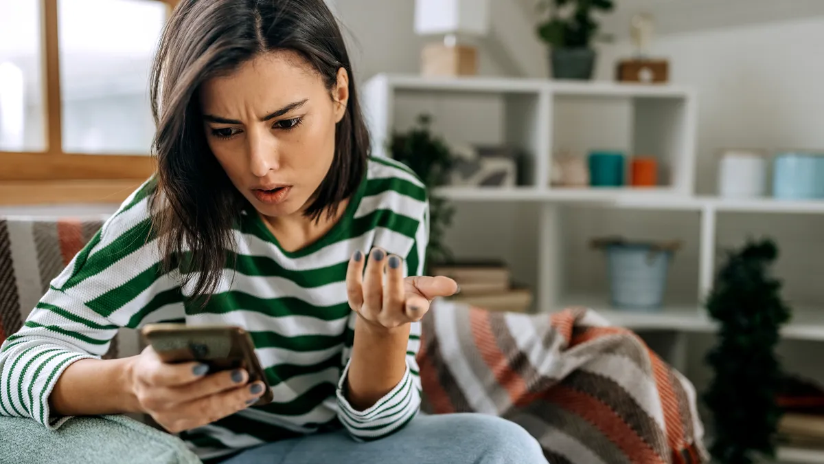 Frustrated young woman with a smartphone in her hands while she sits on a couch at home.