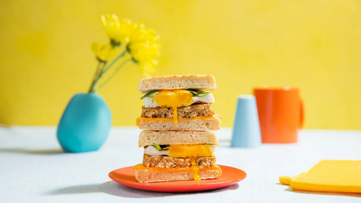Two halves of a breakfast sandwich with cultivated chicken sausage, a poached egg and spinach are stacked on top of each other on an orange plate on a white table with a yellow wall in the background.