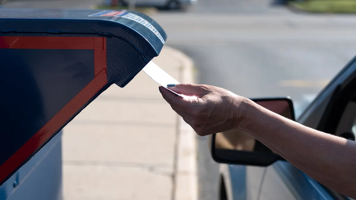 A hand reaches out from a car to deposit a letter in a blue mailbox.