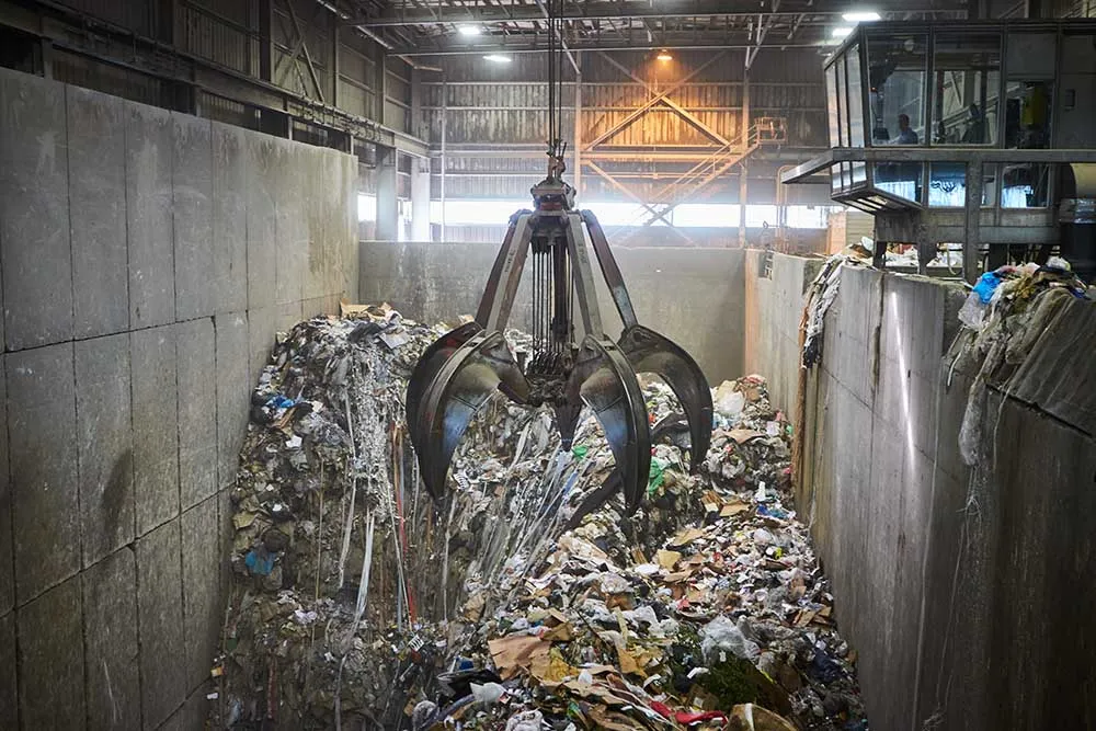 A large claw pulls garbage out of a pile in a concrete pit inside a large building.