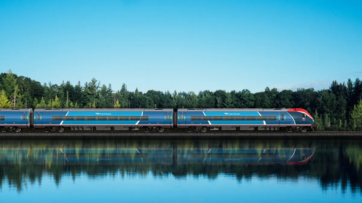 A sleek Amtrak rain painted in two shades of blue accented by red and white is seen along a body of water with green trees in the background and a clear blue sky above.