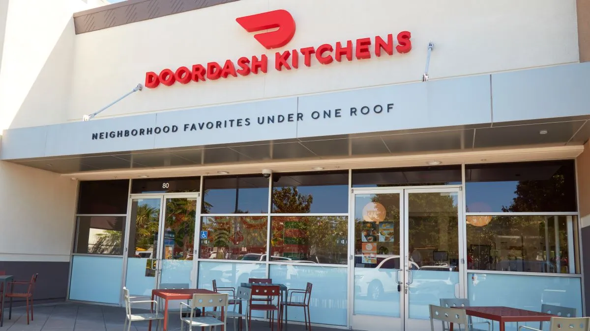 An exterior image of a white building with several tables and chairs. Signage says "DoorDash Kitchens."