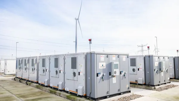 Battery energy storage units at a renewable energy facility with wind turbines in the background.