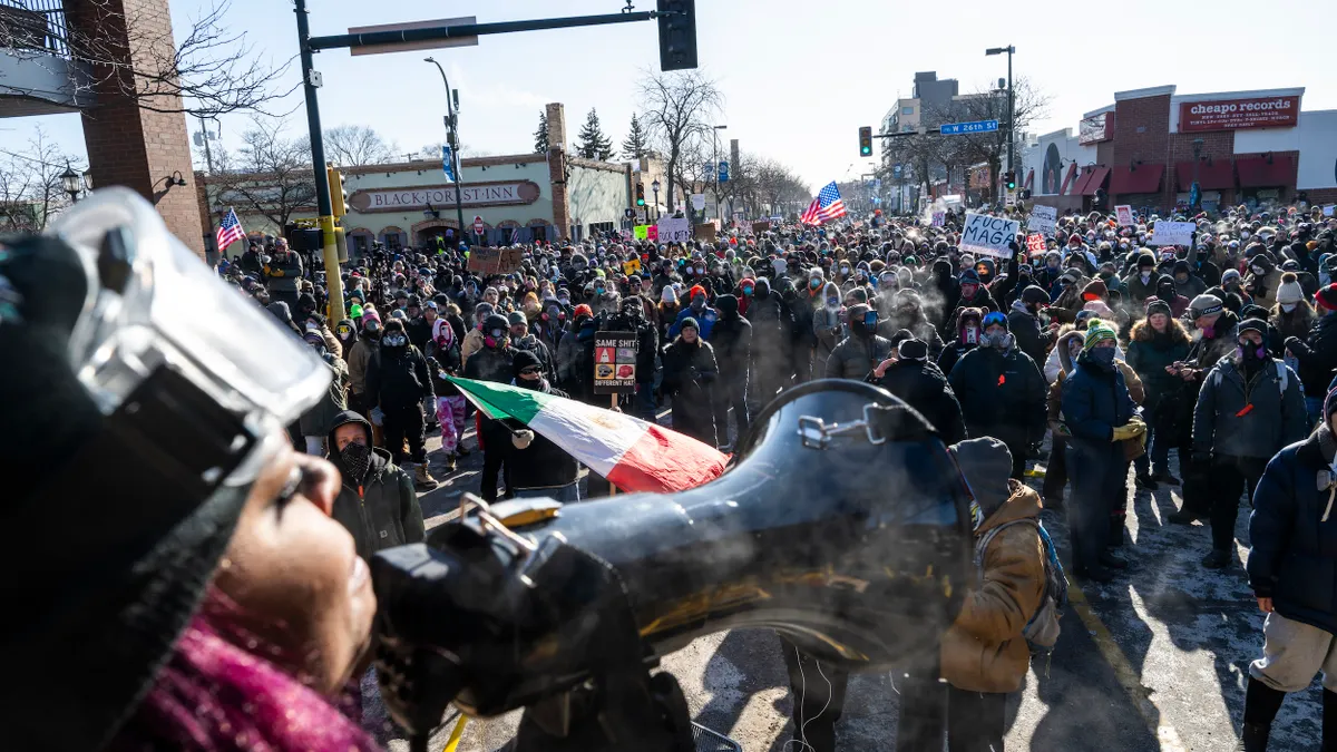 A person wearing a helmet speaks into a bullhorn to a large crowd holding signs.