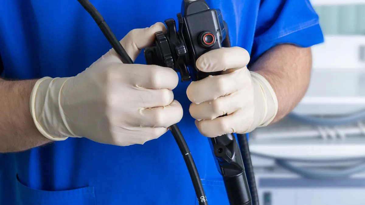 A medical staff member dressed in blue scrubs and with white latex gloves holds a black endoscope