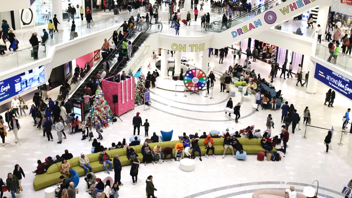 A crowd of shoppers move and mill about in the American Dream mall in East Rutherford, New Jersey. There are people on multiple levels walking around with shopping bags in hand, dressed for fall.