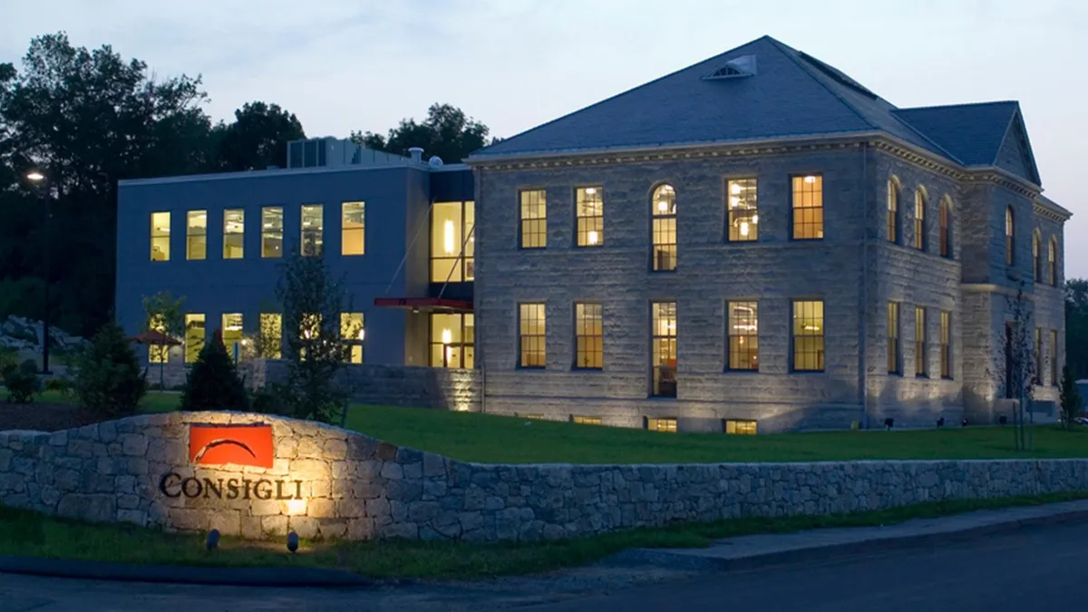 A large stone building looms in the background in a shot at dusk. A sign, with light shining on it, shows Consigli's name.