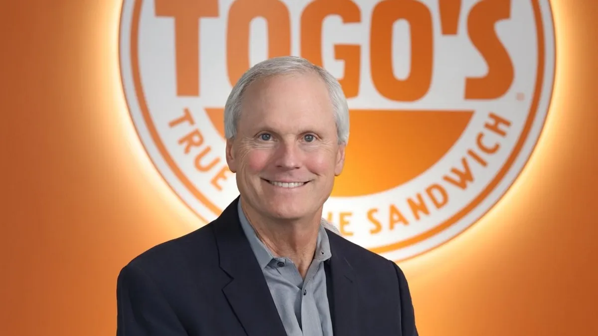 An image of a man wearing a suit in front of an orange logo sign that says "Togo's"