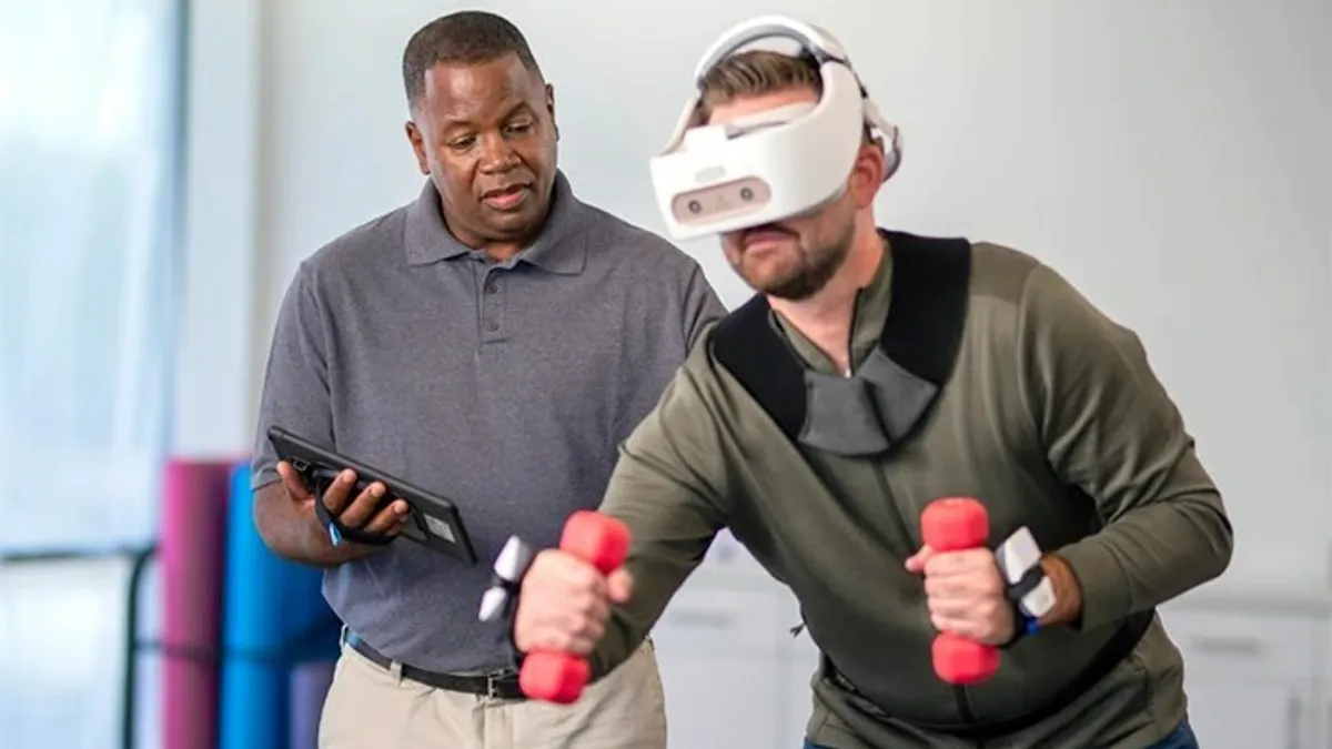 A person holding a tablet watches as another person wears VR googles and does exercises with dumbbells.