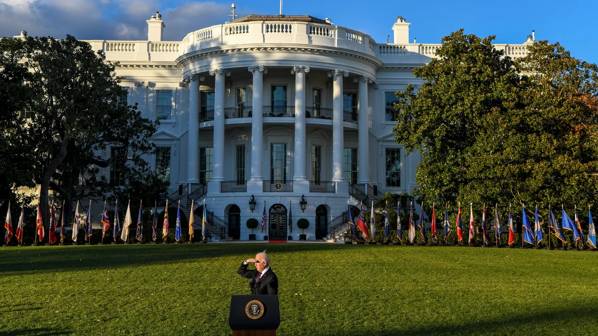 The president joe biden stands behind a podium in front of the white house, shielding his eyes from the sun.