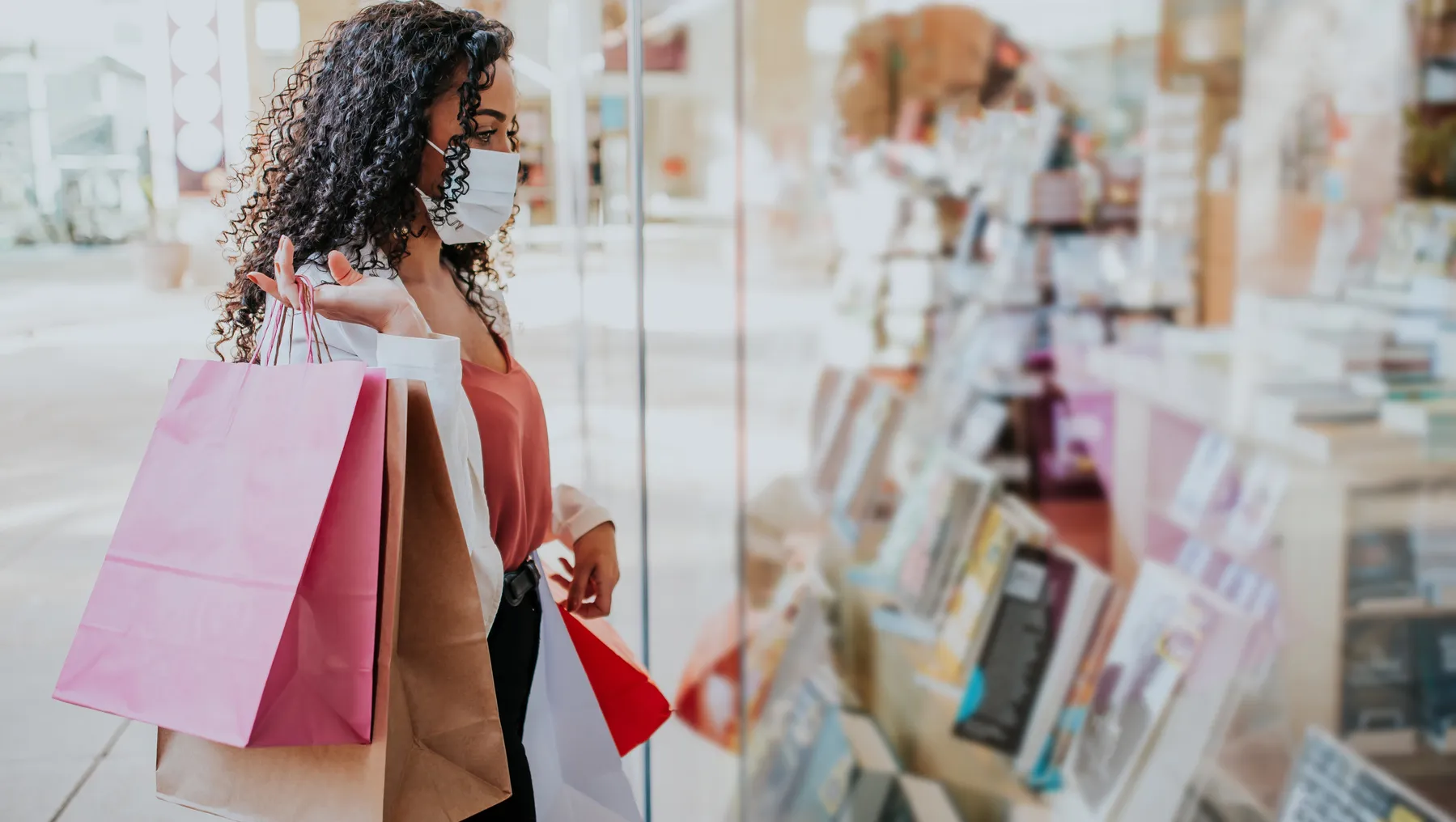 Person with a mask on looks into a store window while holding shopping bags.