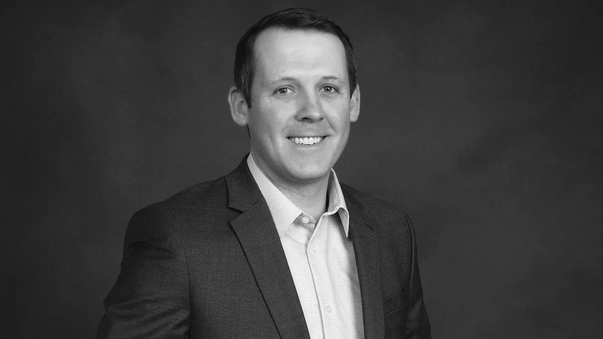 A black and white image of a person wearing a suit and smiling in front of of a gray background.