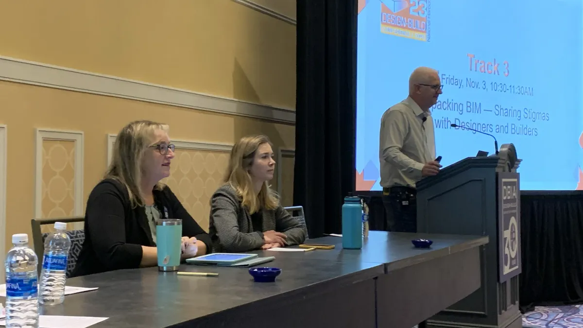 A man with white hair stands at a podium and speaks to a room full of attendees. Two of his fellow panelists in business clothes look on, a slide deck in the background.