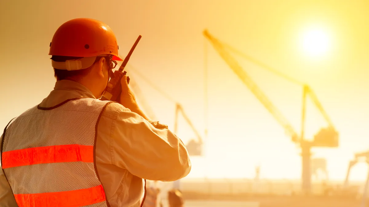 A construction worker speaks into a walkie talkie while under hot sun on a jobsite.