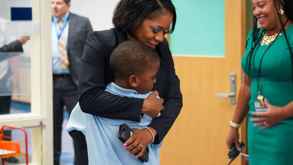 An adult embraces a younger student in a school building while other adult looks on smiling.