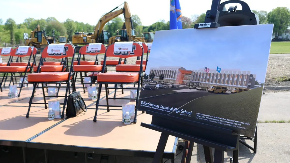 A rendering of a building sits on an easel in front of chairs and construction machinery in the background.