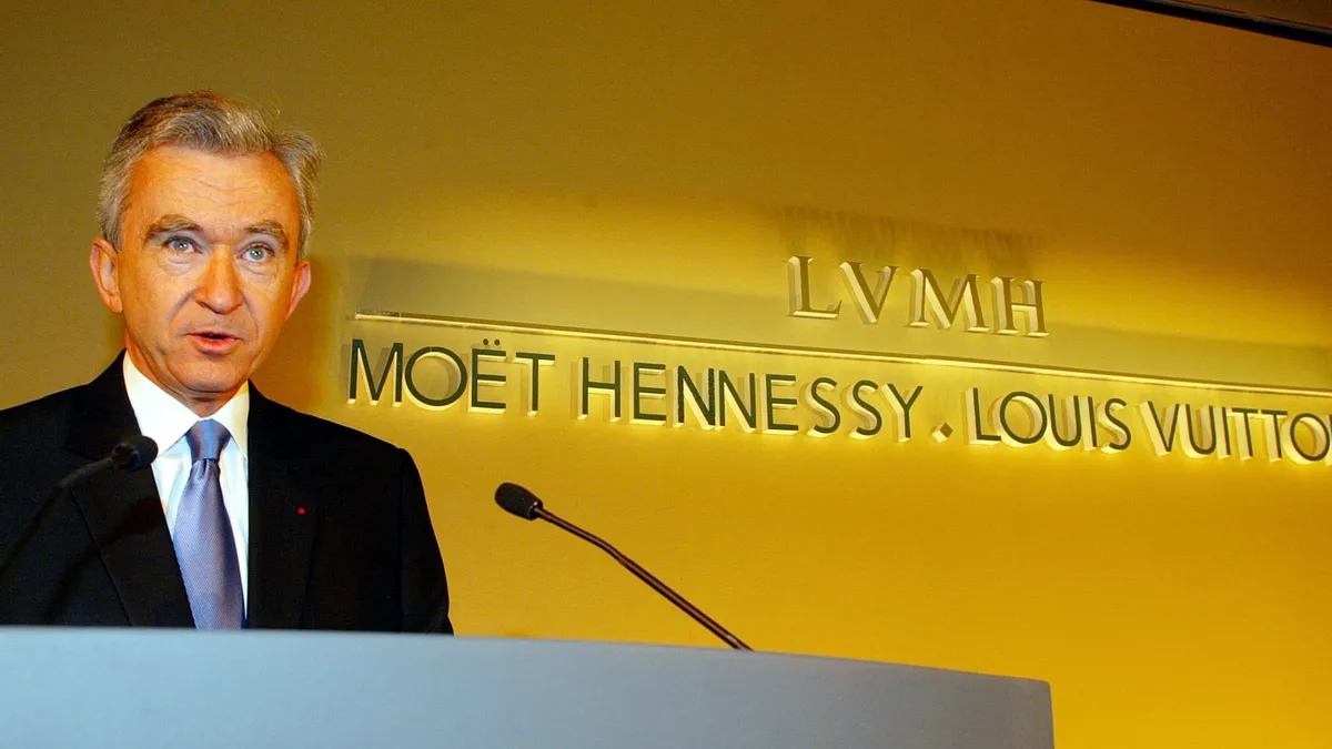 A man in a suit stands in front of a gold wall speaking into a microphone.