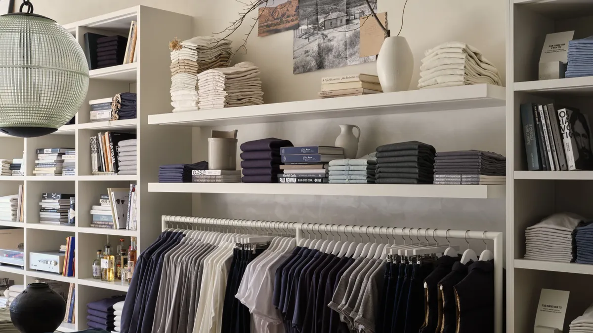 Interior of a Buck Mason store. Shirts and tops are lined up on hangars. Shelves contain folded shirts, white vases and a few books.