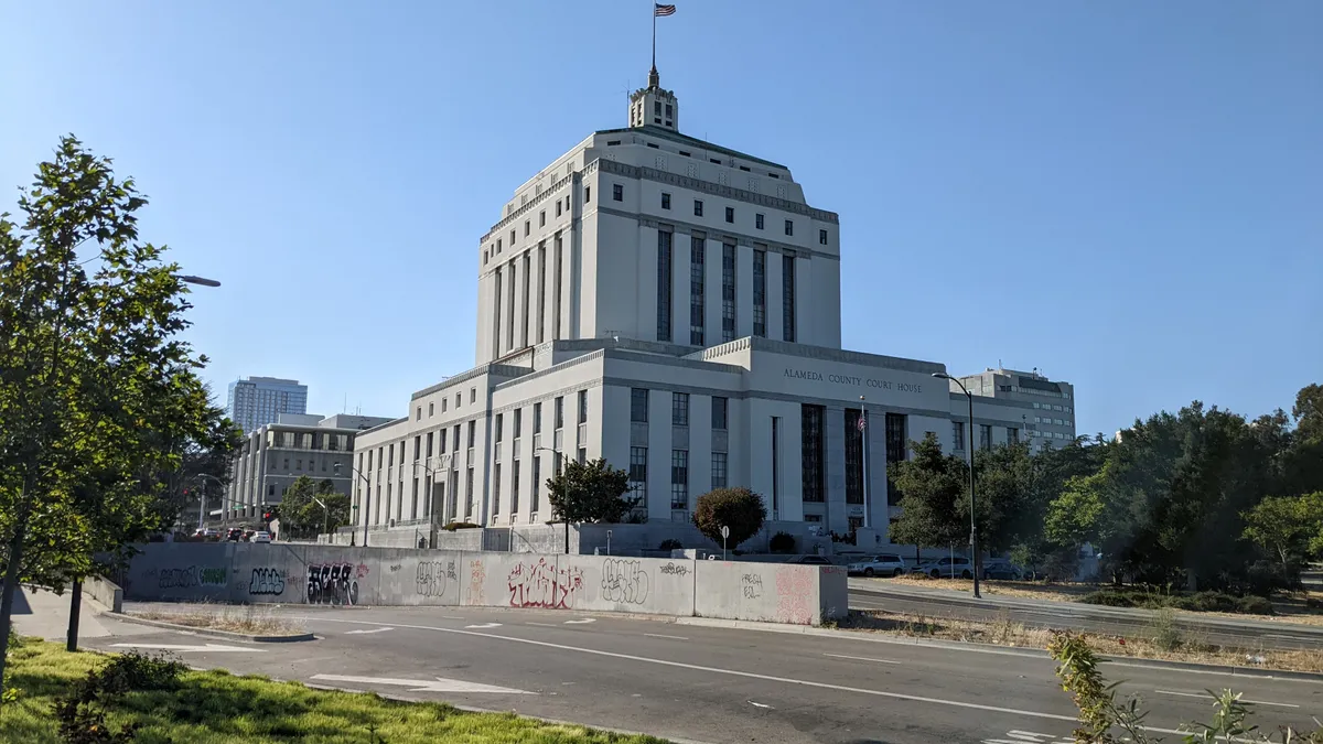 The exterior of a marble government building that is several stories tall crowned by a flag pole, depicted on a sunny day.