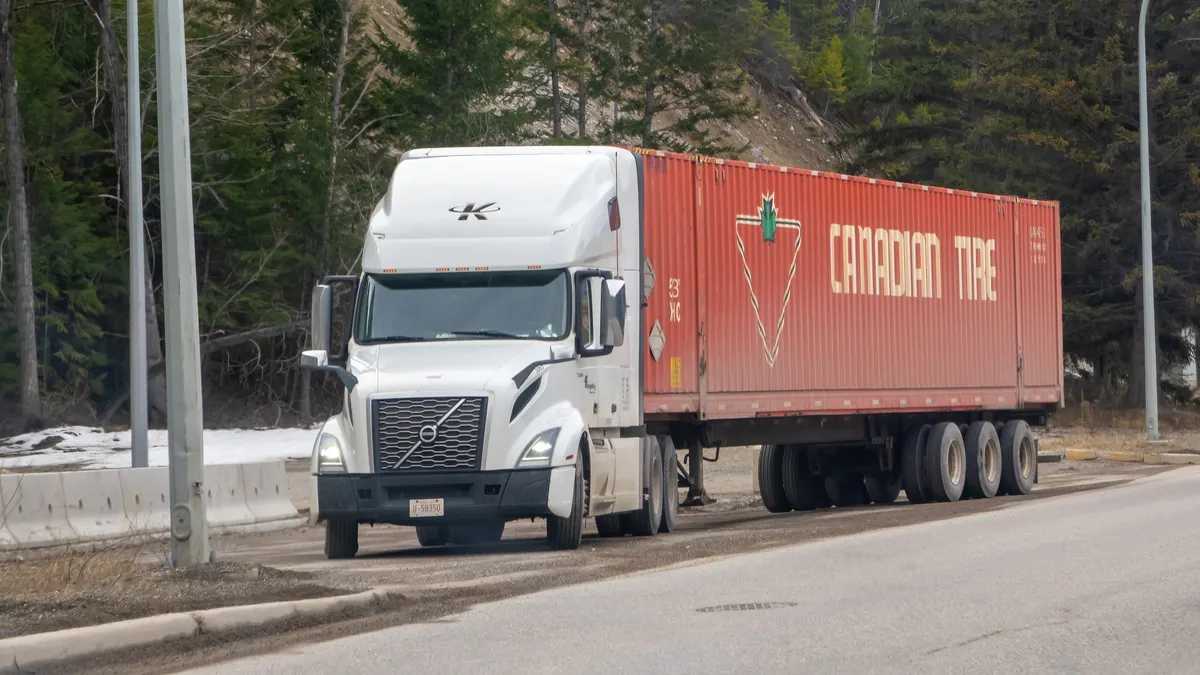 A white Volvo semi-truck with a red Canadian Tire trailer in British Columbia with a wooded hillside in the background.