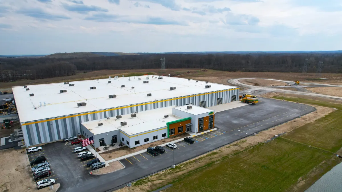 Aerial view of a large, newly built industrial warehouse with a white roof and striped exterior, featuring a smaller office entrance, parked cars, and a U.S. flag, surrounded by open land and construction areas.
