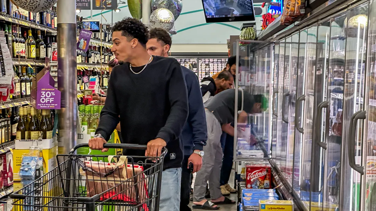 People with a shopping cart walks down a grocery aisle.