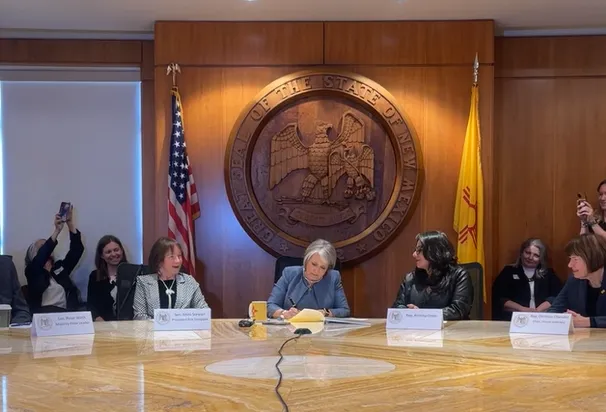 Several people sit at a marble table in front of a large wooden seal of New Mexico. The person sitting center signs a document.