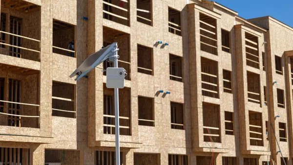 A camera sits on a metal post outside of a brown building on stone amid a bright day.
