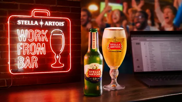A neon red bar sign which reads "Work From Bar" is on a wall. A can and glass of Stella Artois is in the foreground next to a computer while people cheer in the background.