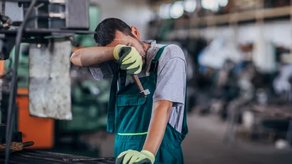 A person wearing coveralls wipes sweat from their in an industrial facility