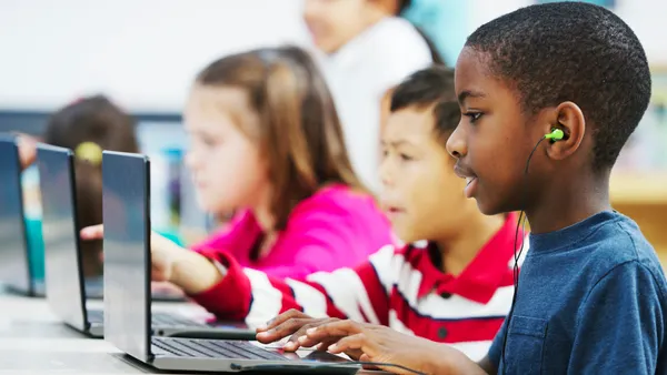 Three younger students sit in a row at a table, In front of them are open laptops. They are typing on keyboards