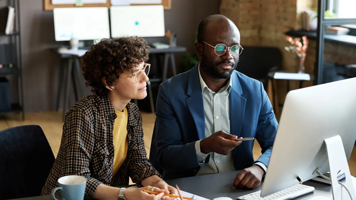 Two colleagues sitting at table in front of computer monitor and discussing work.