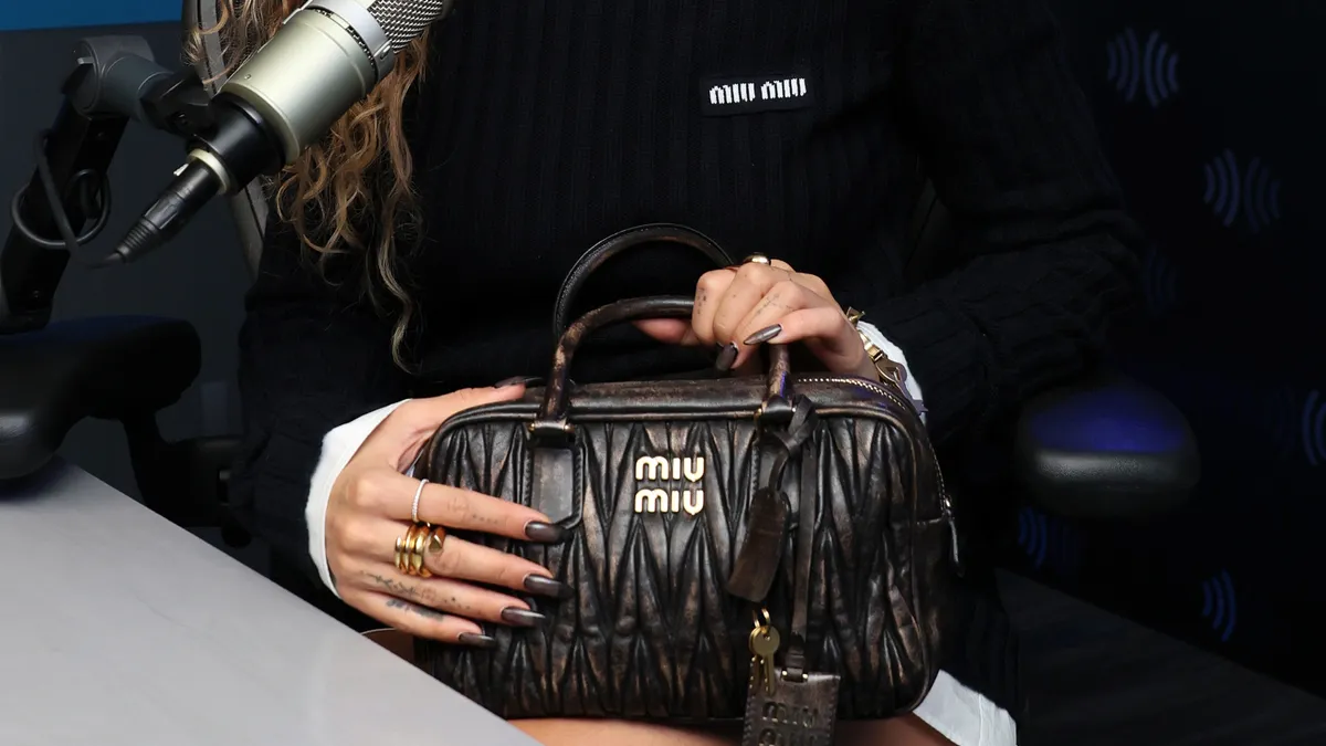 A person sits a table with a microphone holding a Miu Miu handbag and wearing rings.