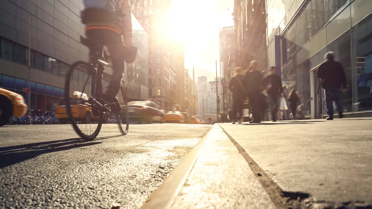 A rider on a bicycle in a bike lane alongside a sidewalk with people and yellow cabs to the left of the bicyclist.