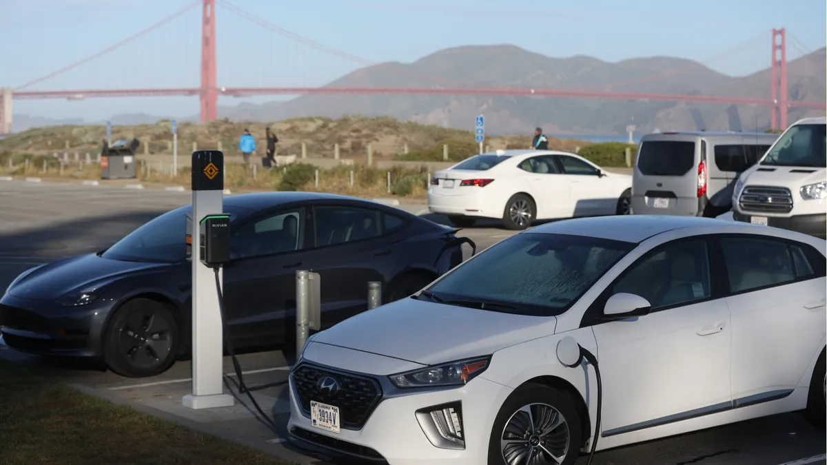 Cars fill a parking lot on a clear, sunny day with people blurred in the background. The cars in the foreground are silver, black, white and more.