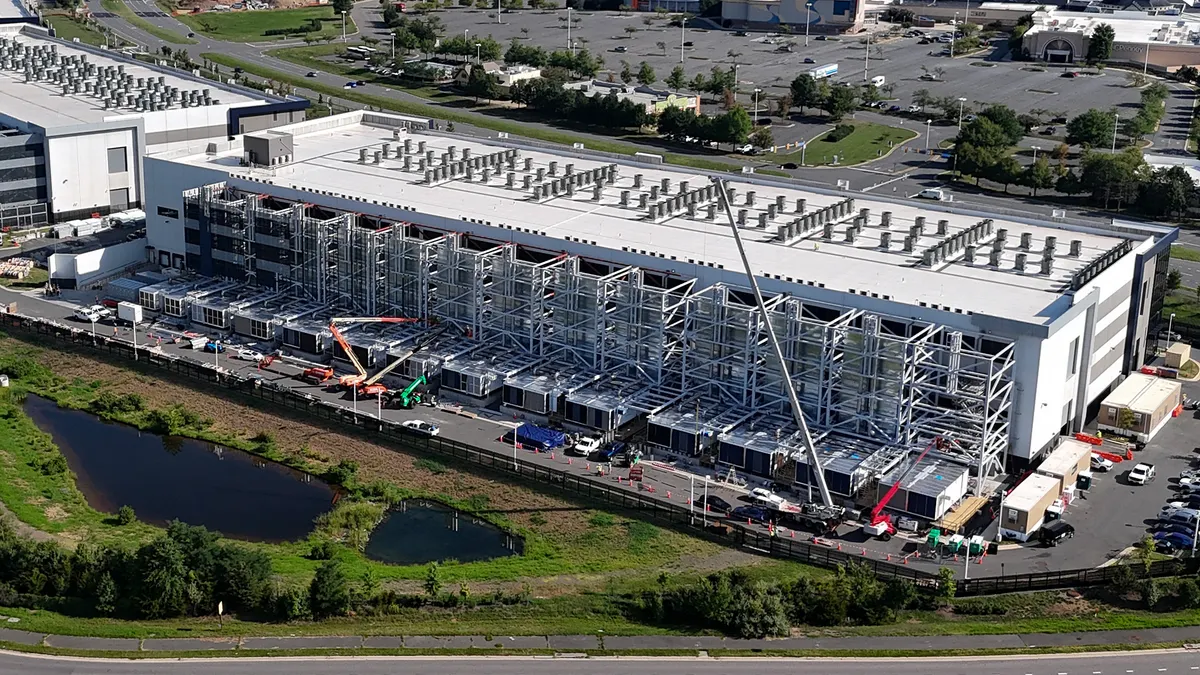 An aerial view of a large data center complex. One multi-story building is complete, while a second, larger building is actively under construction with its steel frame exposed.