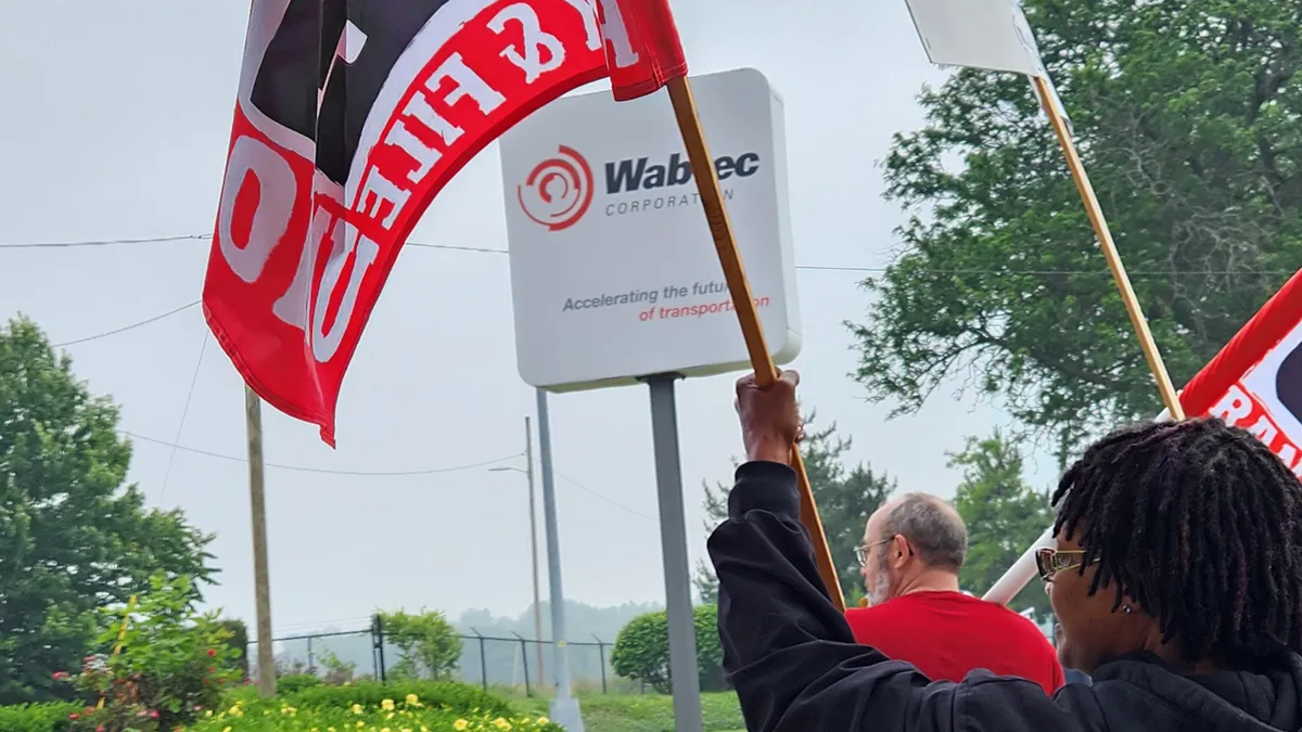 A person waving a red, white and black with the white Wabtec sign in the background.