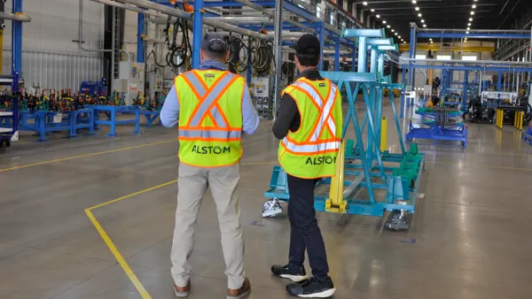 Two men from behind standing on a factory floor wearing visibility vests reading "Alstom."