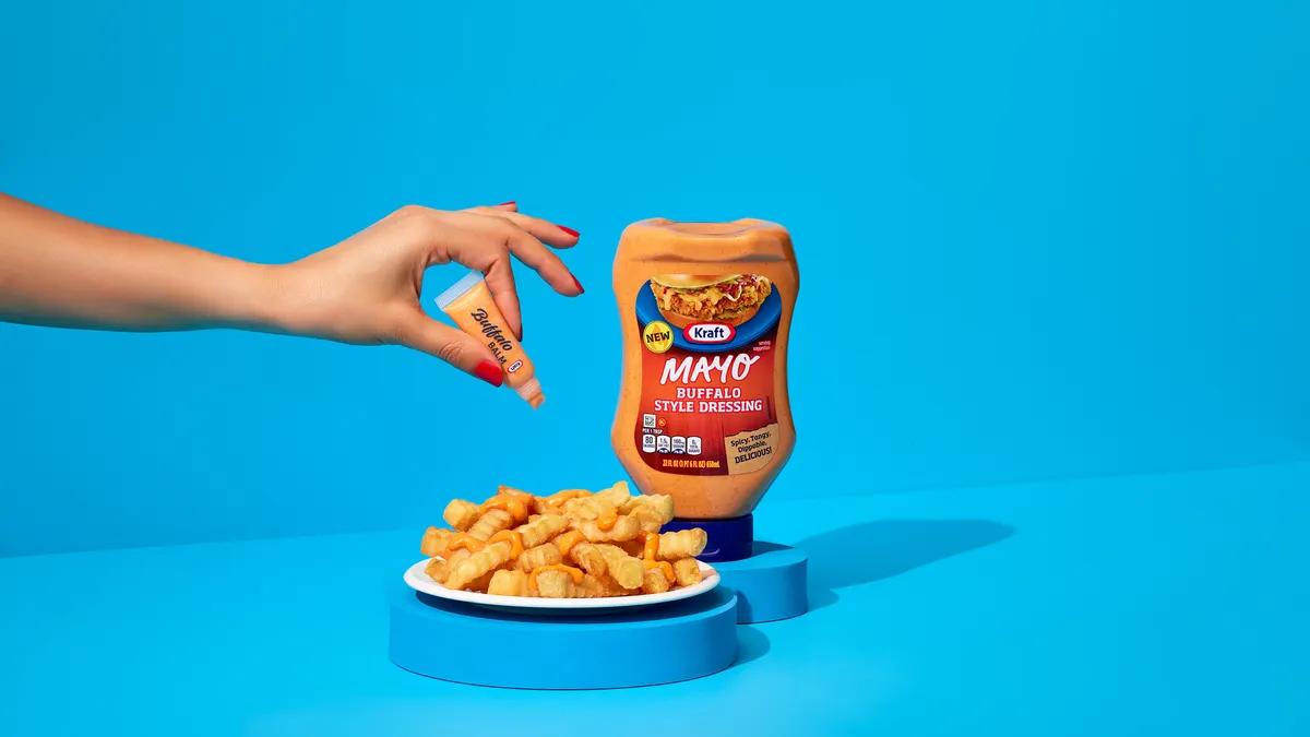 A hand squeezes a small tube of Kraft Mayo Buffalo Style Dressing on a plate of fries. The full sized bottle is to the right of the plate. The background is blue.