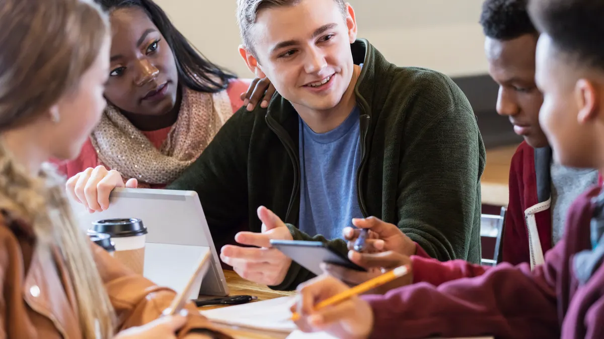 A small multi-ethnic group of teenager high school students sitting at a table in a coffee shop, studying together, using digital tablets and spiral notebooks.
