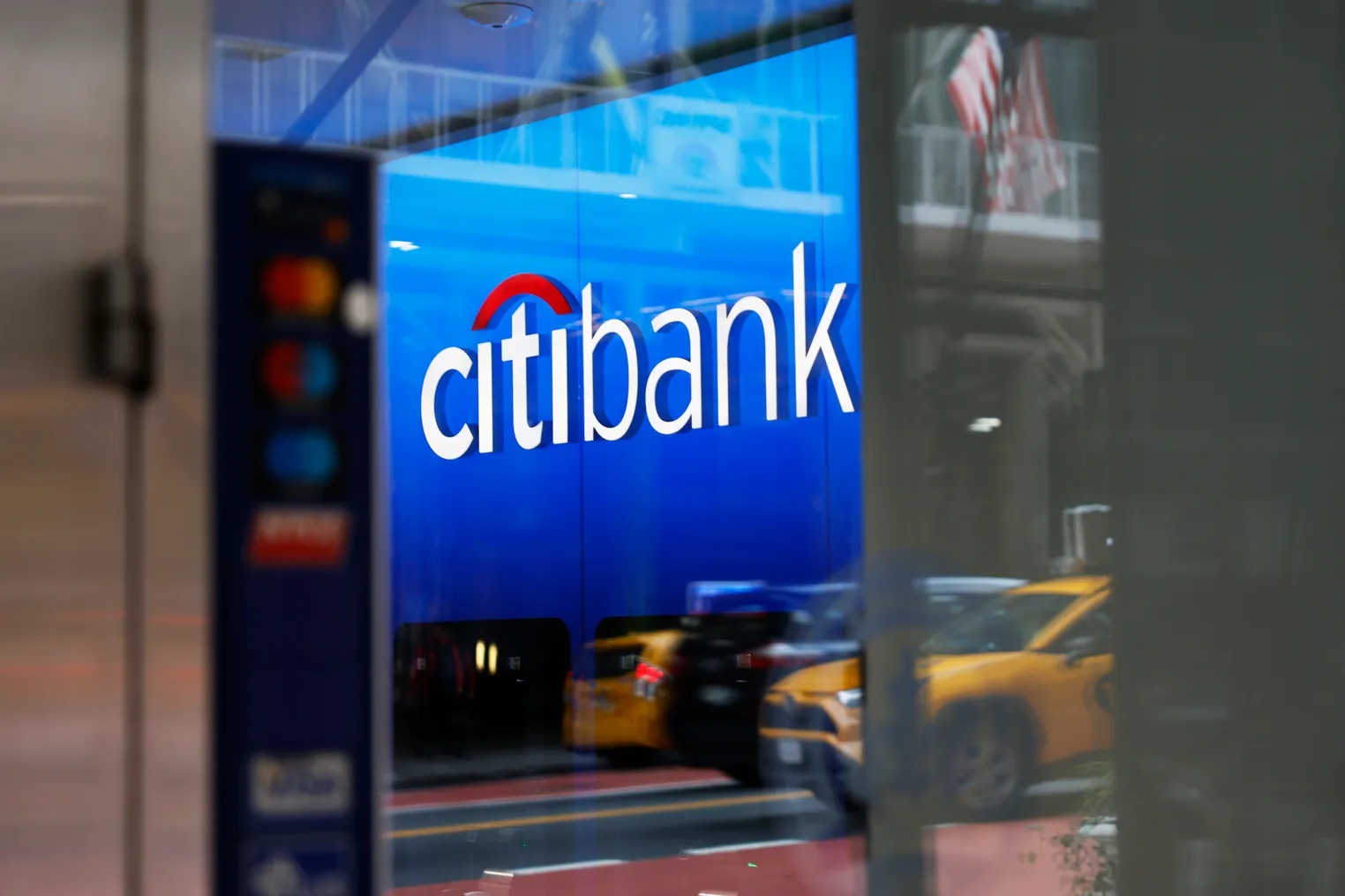 A sign reads "Citibank" in a branch window, which reflects an American flag and taxi cabs on the street.