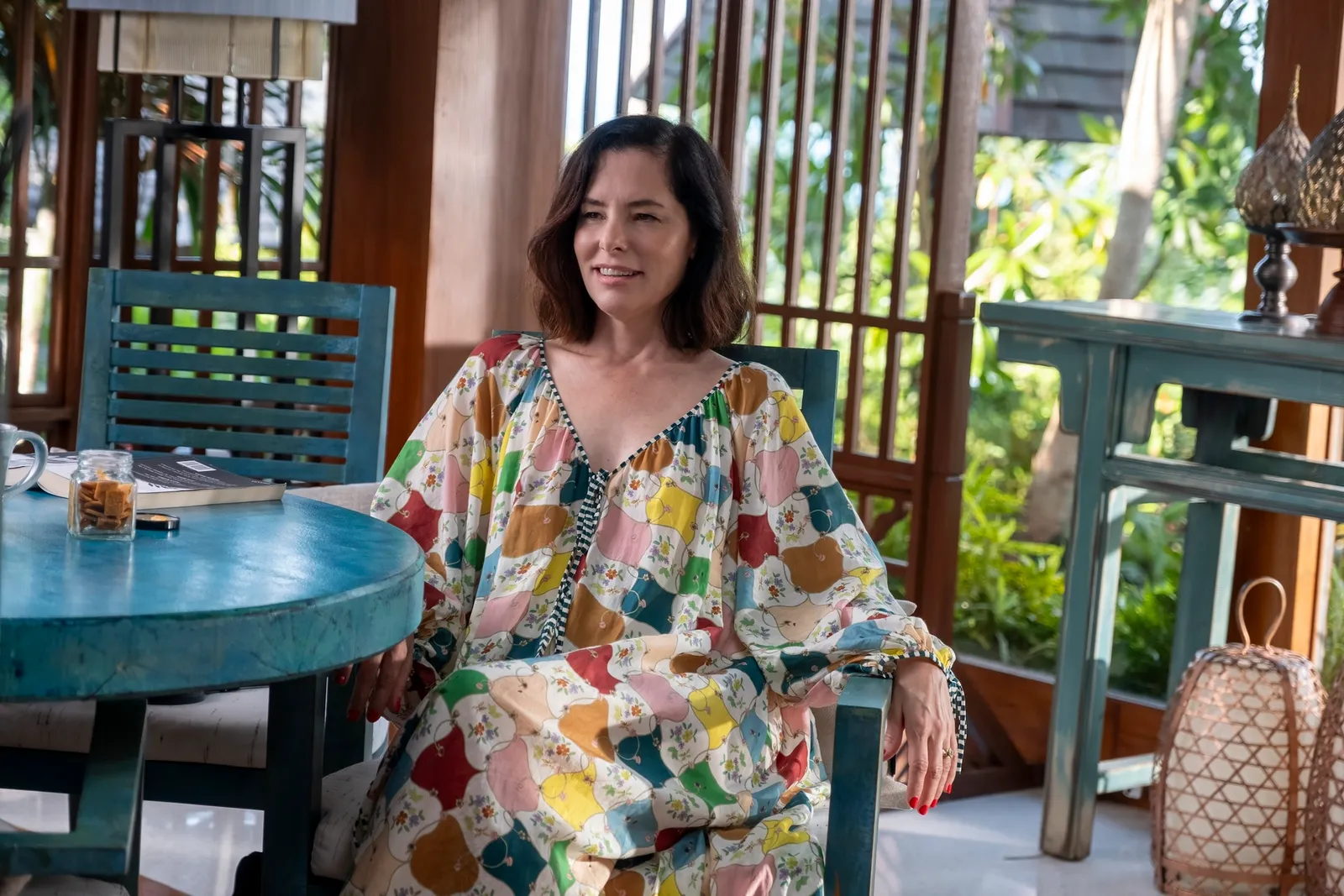 A woman in a colorful flowing dress sits in a blue chair at a blue table on an outdoor patio.