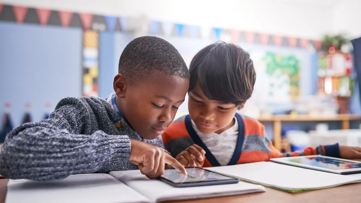 Two elementary students sit together working on a classroom assignment on a tablet.