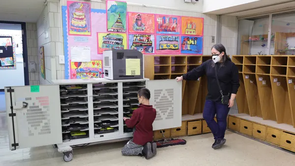 A student is near a cabinet holding laptops. A teacher stands nearby watching the student