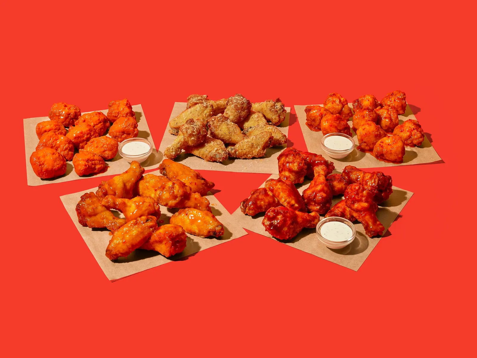 A photo of five different flavors of wings sitting on squares of brown paper against a red background. There are small cups of ranch dipping sauce near three of the types of wings.