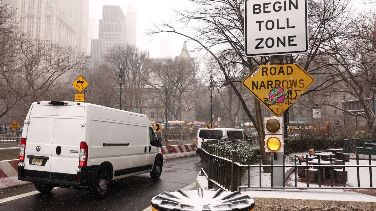 White vans pass a sign that reads, "Begin Toll Zone" and "Road Narrows."