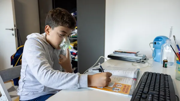 A student is sitting at a desk with a computer and notepad. The student is wearing a oxygen mask that is connected to a nebulizer on the desk.