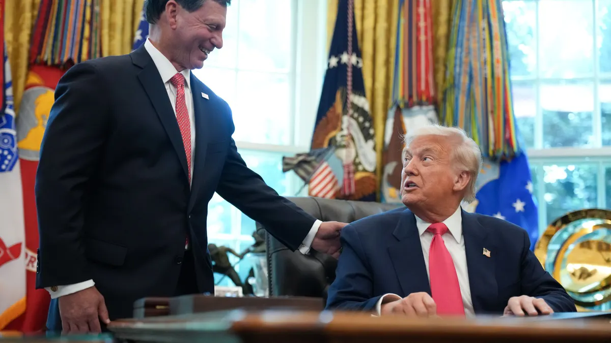 Former Fiserv CEO Frank Bisignano lays hand on shoulder of President Donald Trump in Oval Office with flag in background.