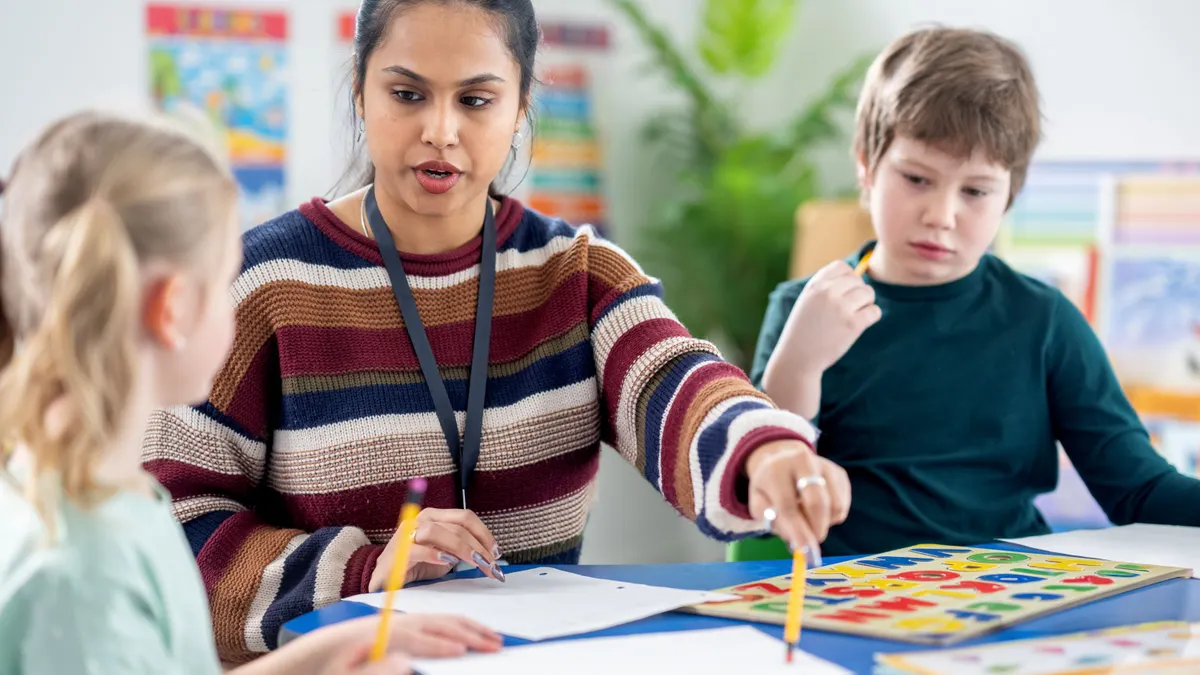 A teacher sits down with two elementary school students while working through a colorful puzzle and worksheet activity at a classroom table.